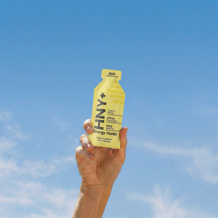 Hand holding an HNY+ Buzz energy honey gel against blue sky
