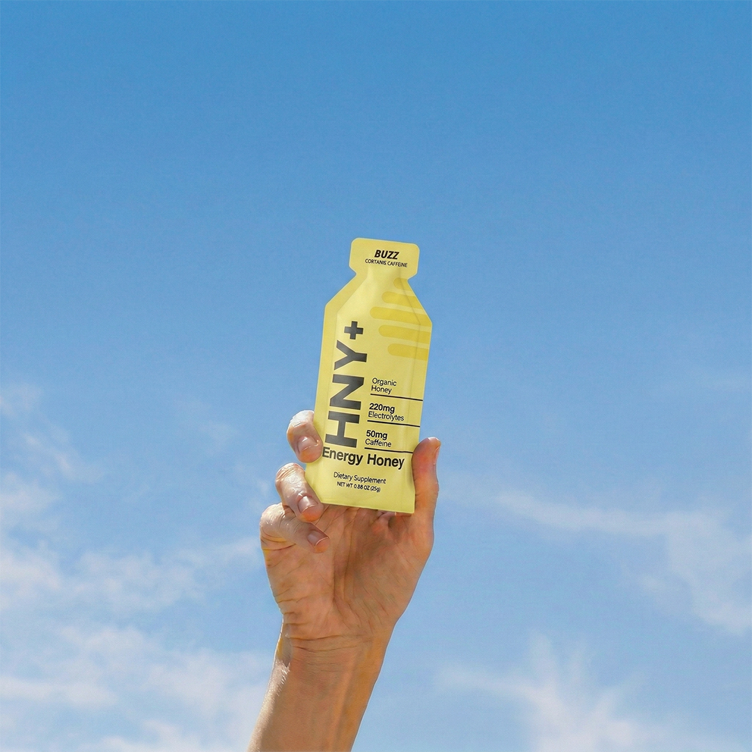 Hand holding an HNY+ Buzz energy honey gel against blue sky