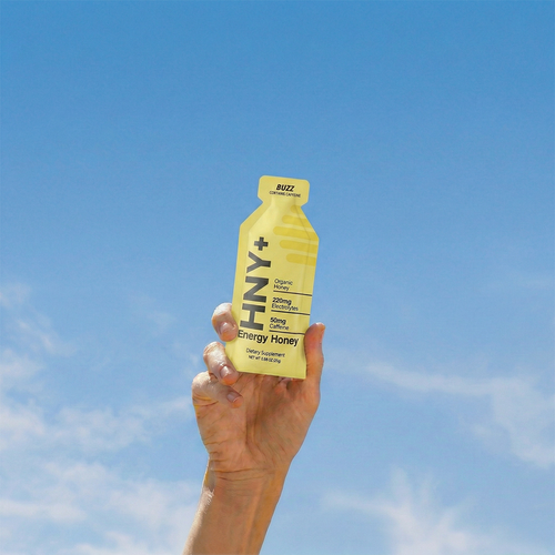 Hand holding an HNY+ Buzz energy honey gel against blue sky