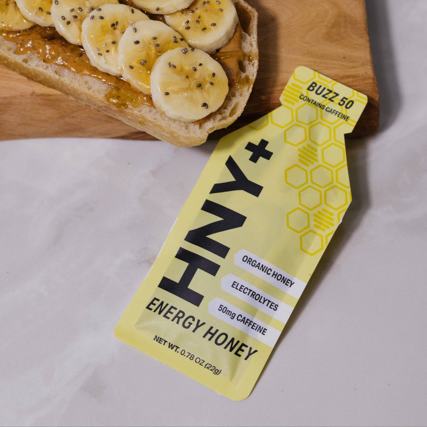 Honey packet labeled 'HNY+' next to a slice of bread with banana slices and peanut butter on a wooden board.